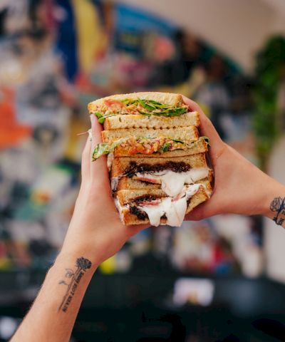 A stacked, gooey sandwich with melted cheese and greens, held up proudly in front of a colorful, blurred backdrop.
