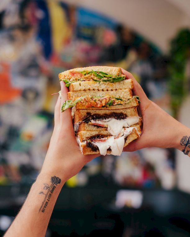A stacked, gooey sandwich with melted cheese and greens, held up proudly in front of a colorful, blurred backdrop.