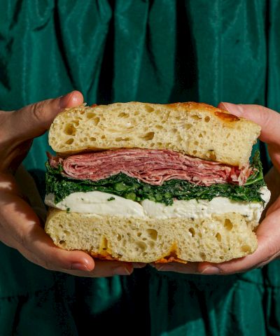 A large sandwich with crusty bread, layers of salami, greens, and cheese, held by hands against a dark green background.