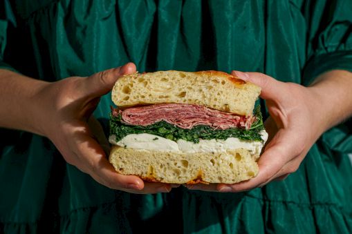 A large sandwich with crusty bread, layers of salami, greens, and cheese, held by hands against a dark green background.