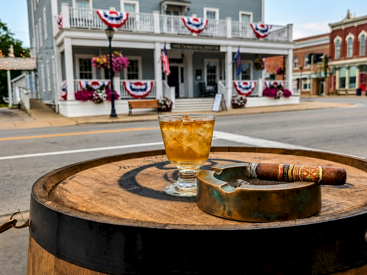 A sunny street patio with an amber drink and cigar on a barrel table, facing a historic building decorated with patriotic bunting.