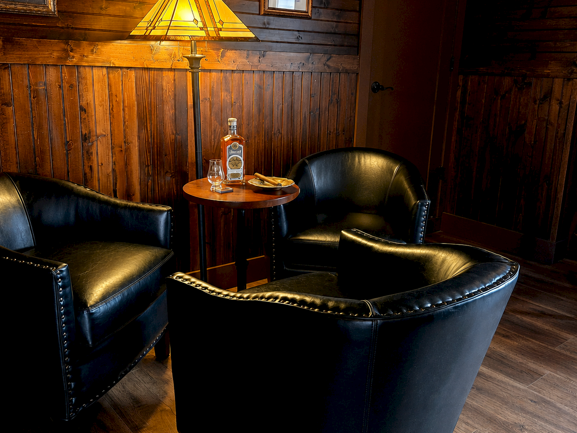 A cozy wood-paneled lounge with leather chairs, a small round table, a stained-glass lamp, and cigar-themed framed posters and decor.